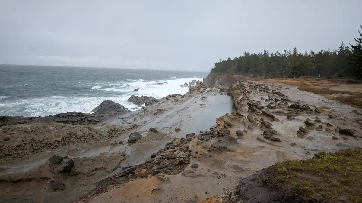 A wide-angle photo of a rugged, wet coastal landscape with layered rock formations, crashing ocean waves, and a distant…