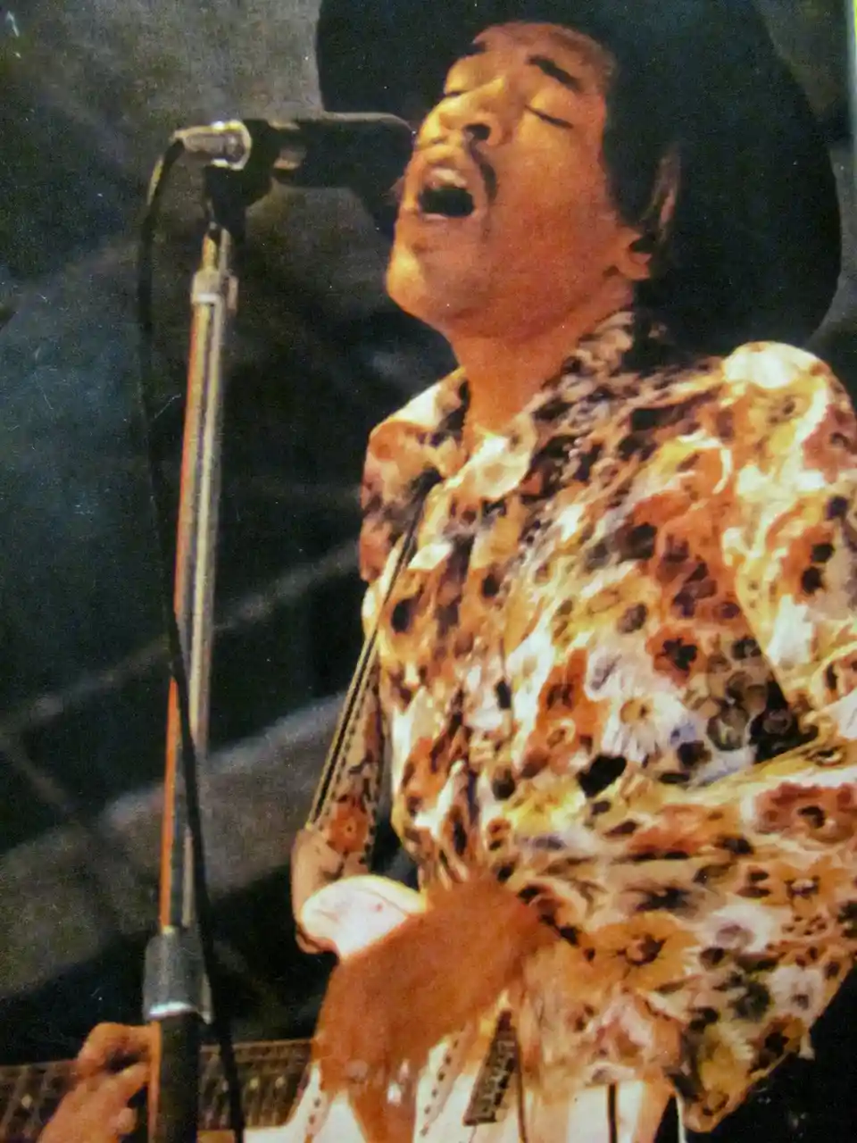 A vintage photo of Jimi Hendrix performing on stage, his head tilted back and mouth open as if screaming into a microphone…
