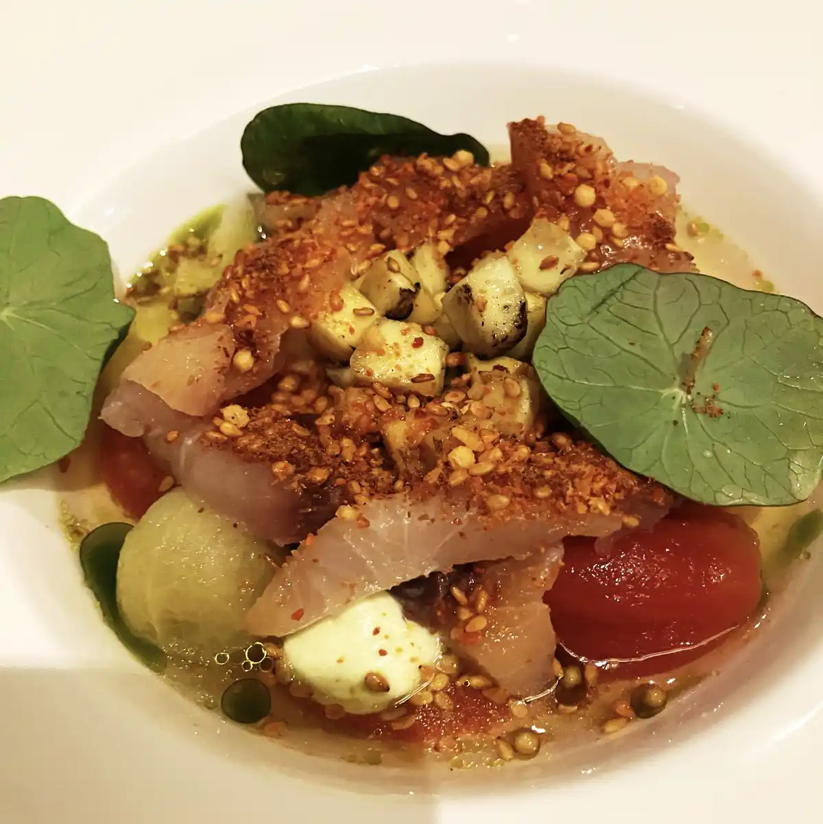 A close-up of a white bowl containing a gourmet dish with translucent fish slices, toasted sesame seeds, small cubes of what…
