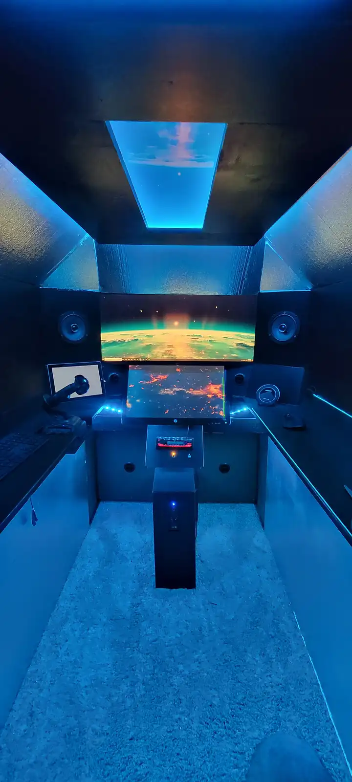A custom-built, narrow, blue-lit room resembling a spaceship cockpit, featuring multiple monitors displaying space scenes…