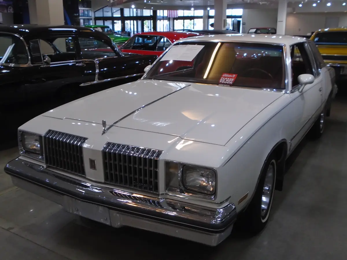 A white 1970s Oldsmobile sedan is displayed in a car museum, with a red 'PLEASE DO NOT TOUCH' sticker visible on its…