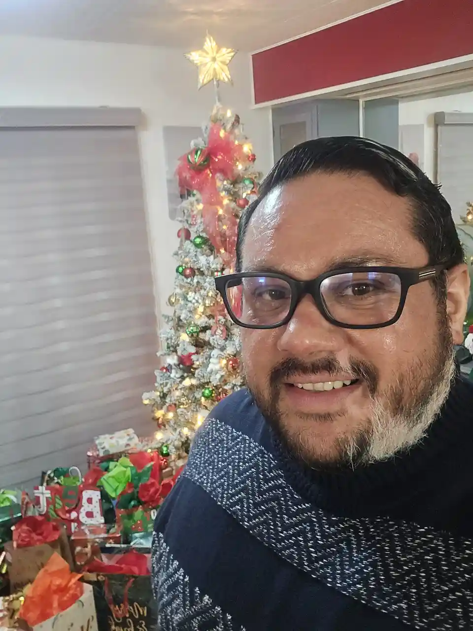 A man with glasses and a beard smiles at the camera, standing in front of a decorated Christmas tree with lights and…