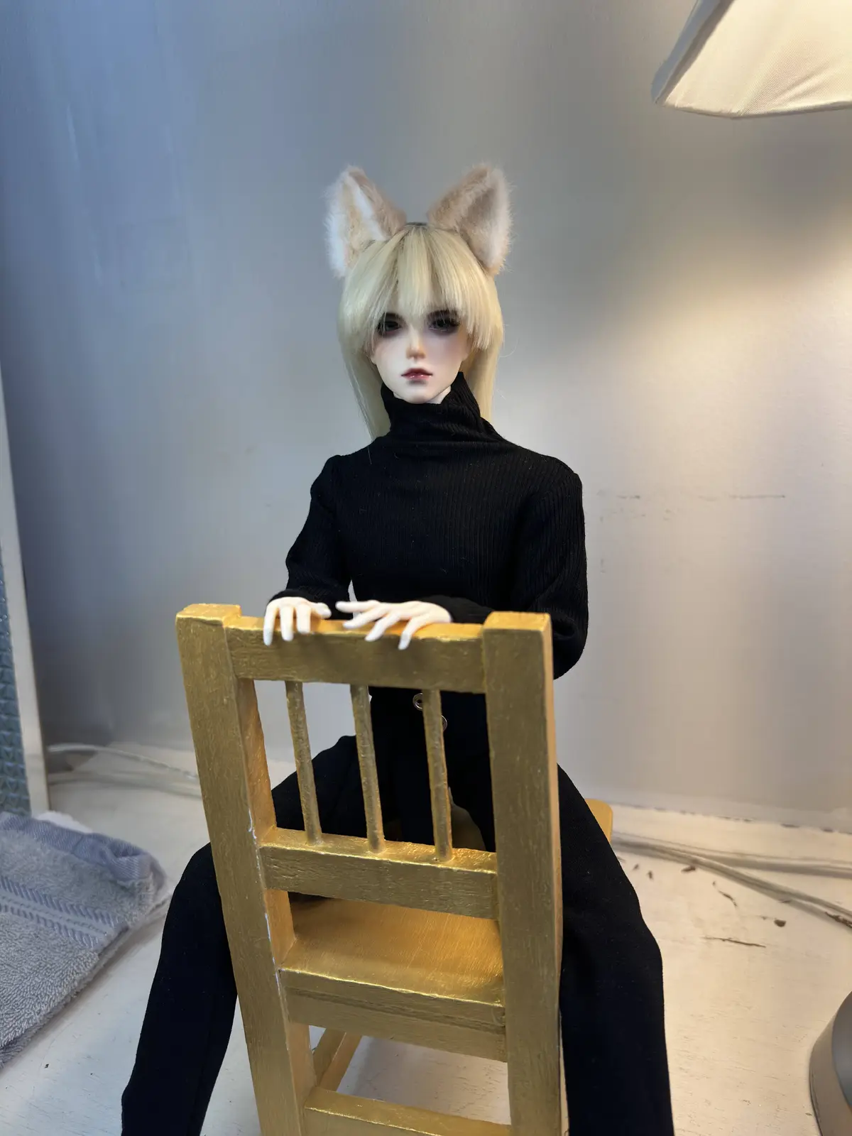 A doll with blonde hair and cat ears sits on a gold-painted wooden chair, wearing a black turtleneck and black pants, with…