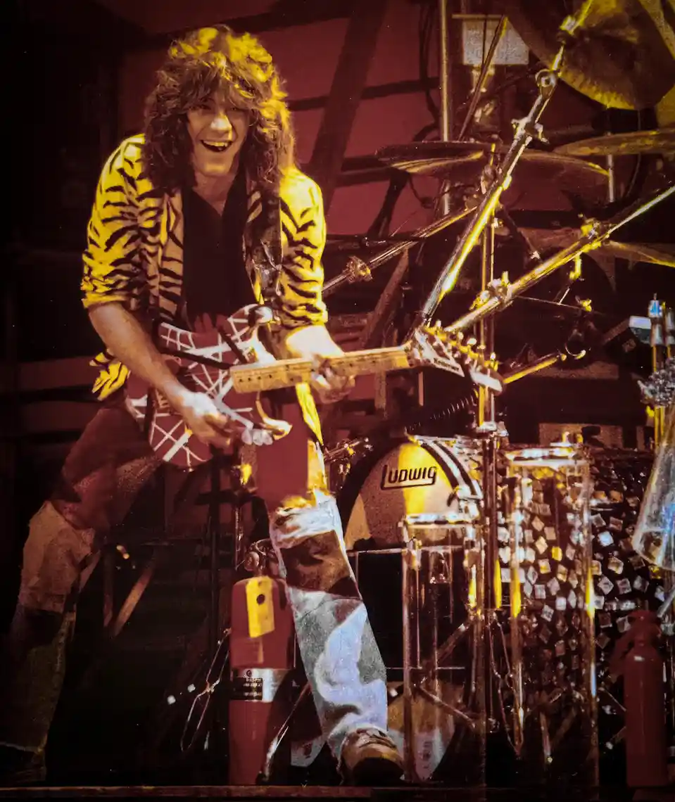A man with long curly hair, wearing a tiger-print shirt and ripped jeans, is smiling while playing a red and white electric…