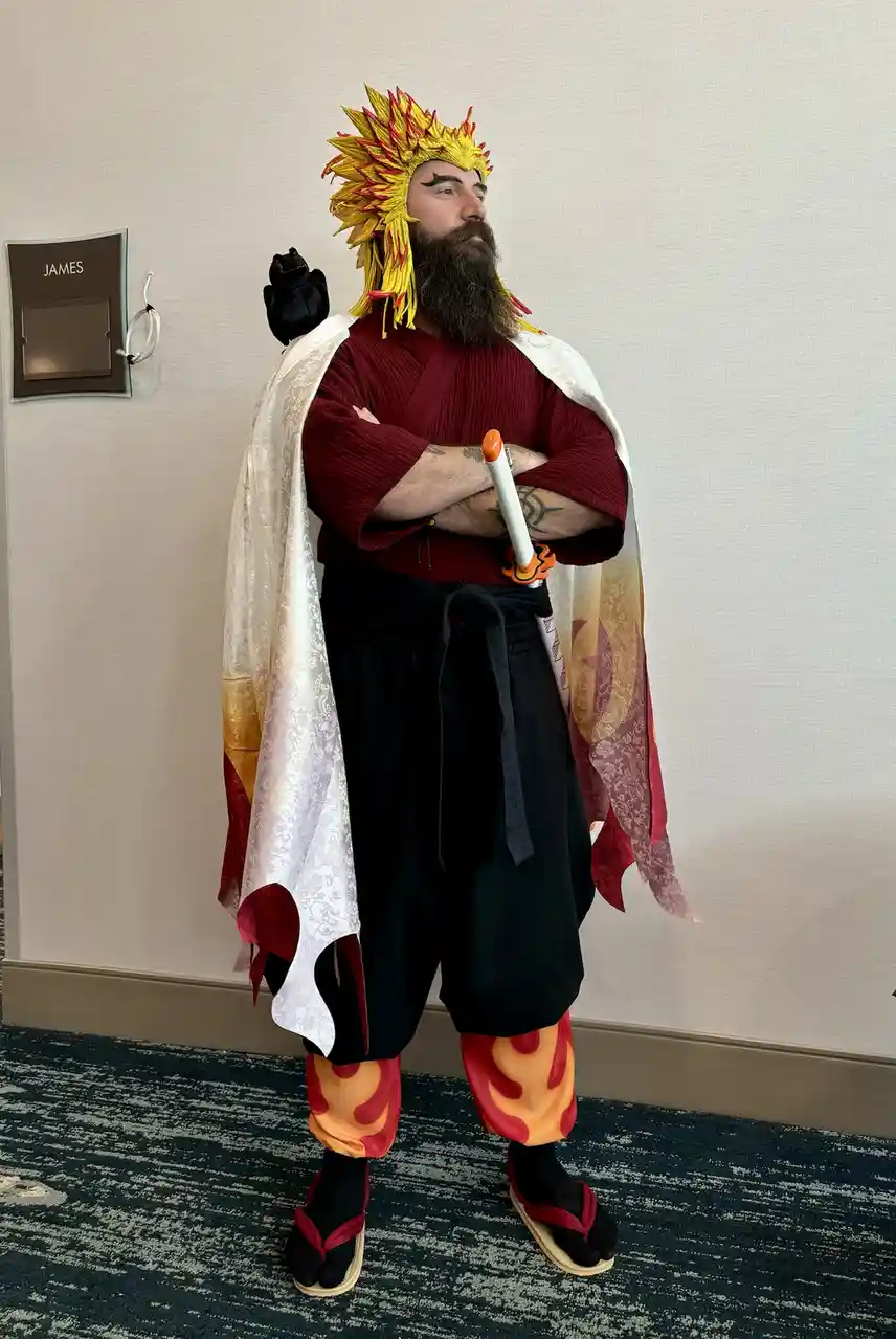 A man with a long beard and dramatic makeup stands confidently in a full cosplay outfit resembling a fiery anime character…