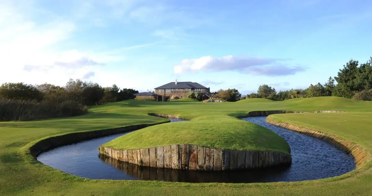 A scenic golf course photo shows a winding water hazard surrounding a grassy island with a house in the background. The…