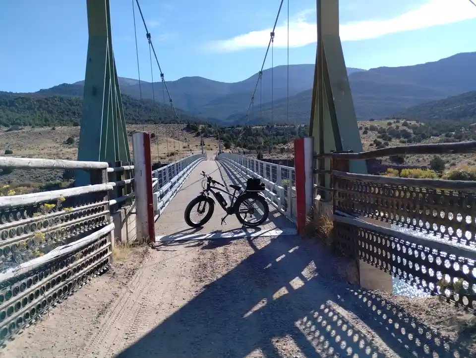 A black electric bicycle with a rear rack and bag is parked on the dirt path at the entrance of a metal suspension bridge…