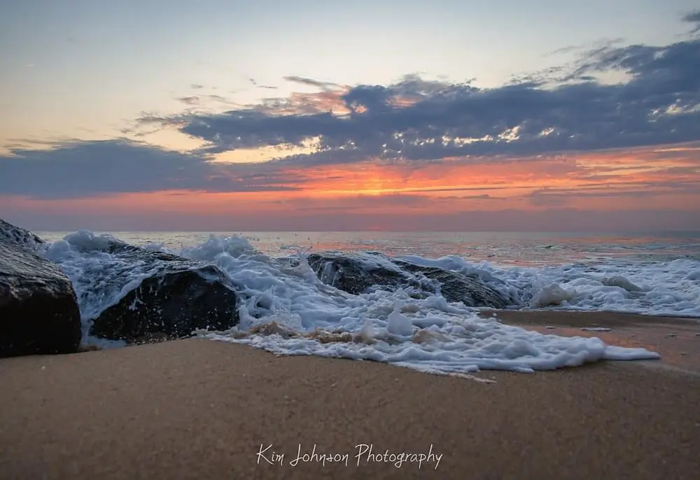 A serene beach scene at sunset with waves crashing over dark rocks onto sandy shore, sky painted with orange, pink, and…