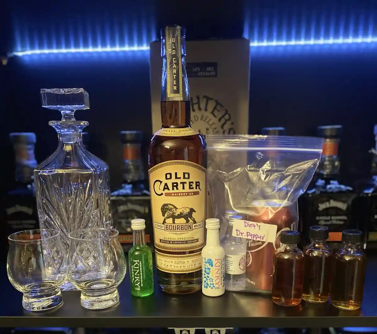 A collection of whiskey bottles, glasses, and small flavoring bottles arranged on a shelf with blue backlighting. A large…