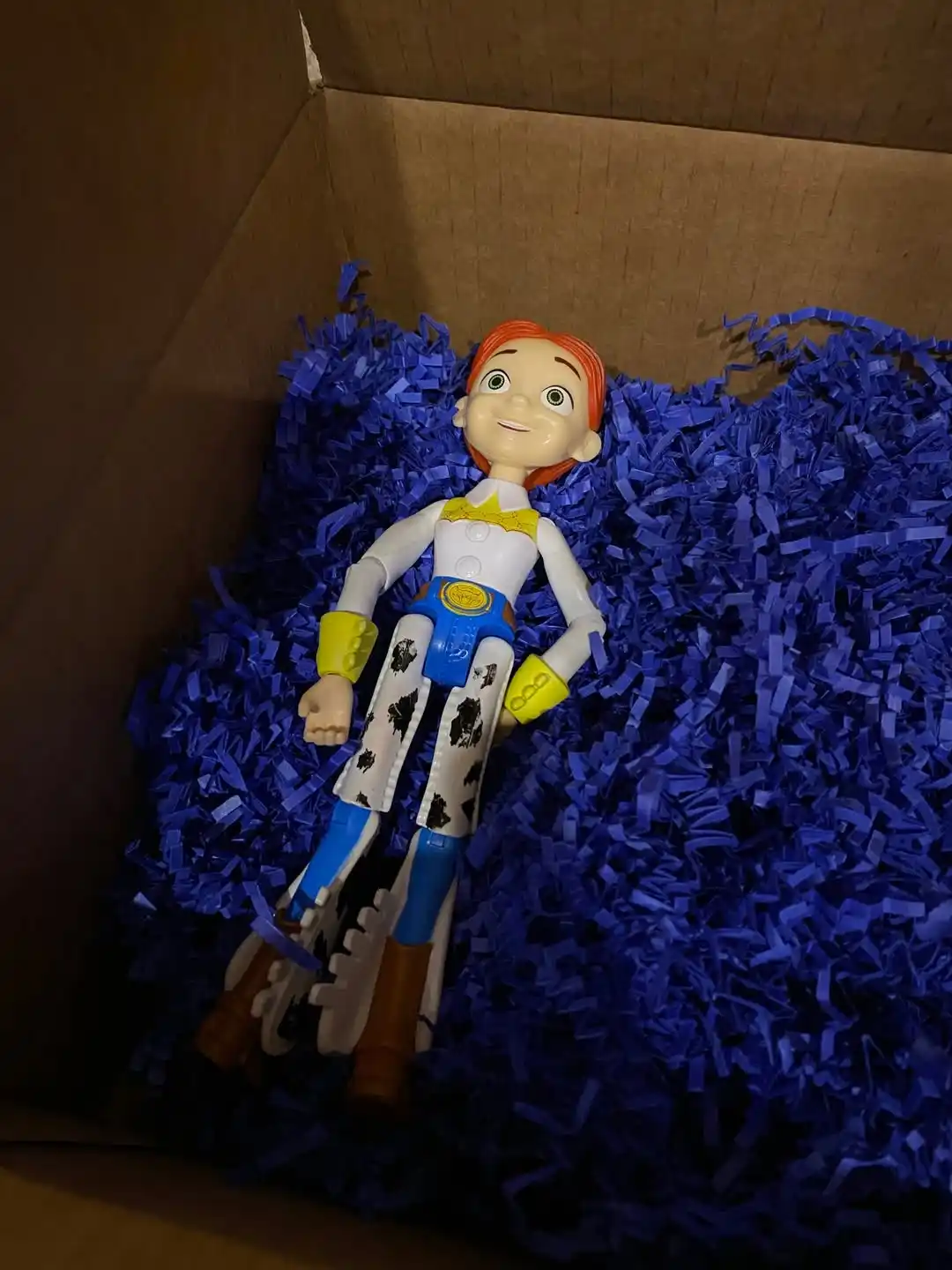 A toy doll resembling Jessie from Toy Story lies in a cardboard box filled with blue shredded paper. The doll has orange…