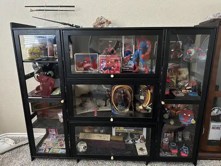 A black glass-front display cabinet is filled with various Spider-Man merchandise, including action figures, lunchboxes, and…