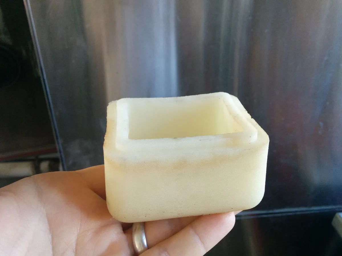 A hand holds a pale, rectangular, 3d-printed object with a hollow center and visible layer lines, resembling a soap bar but…
