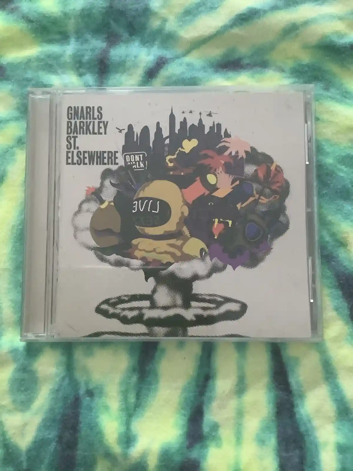 A CD case for Gnarls Barkley's album 'St. Elsewhere' rests on a tie-dye fabric. The cover art features a stylized, chaotic…