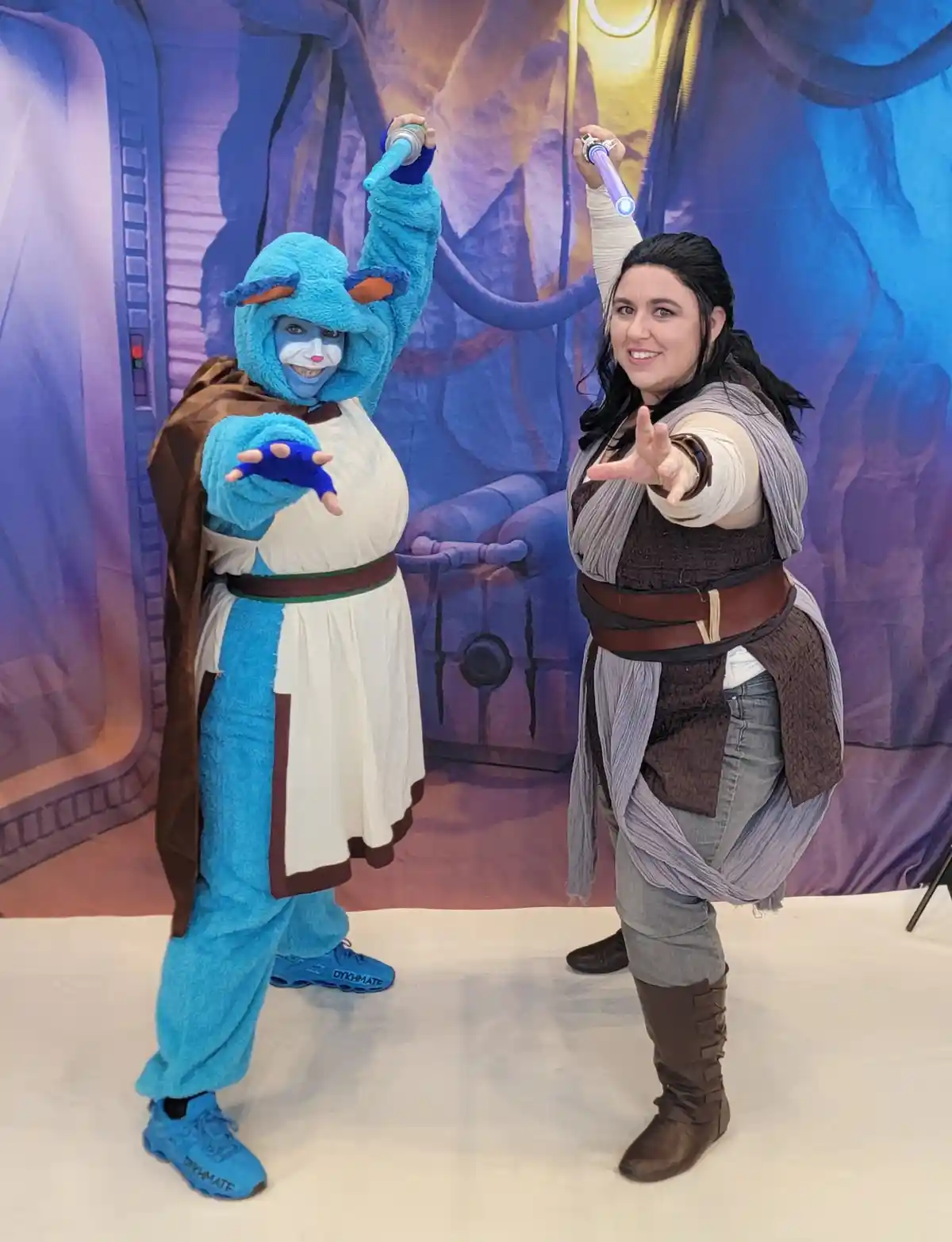 Two people pose in front of a Star Wars-themed backdrop, one dressed as a blue furry creature in Jedi robes holding a blue…