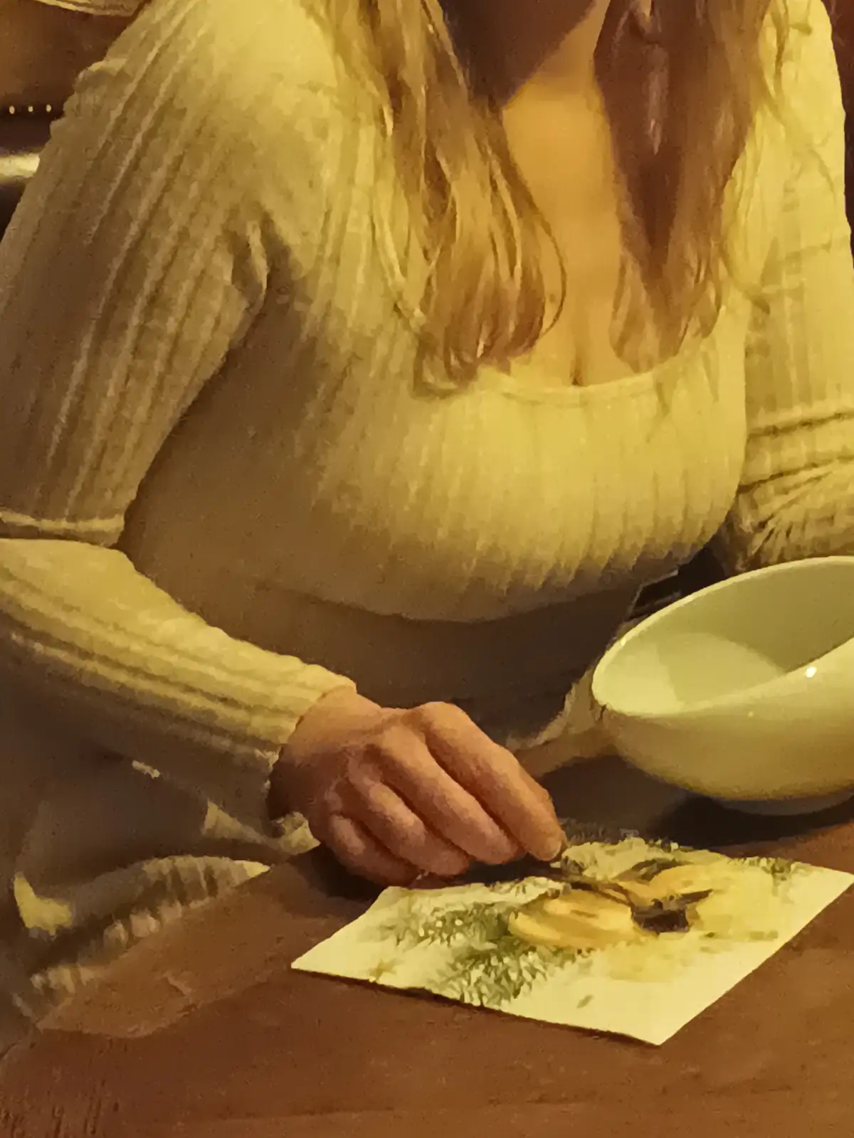 A person in a yellow ribbed sweater is seated at a wooden table, using a brush to paint on a small piece of paper. A white…