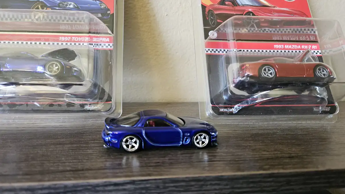 A blue die-cast toy car sits on a wooden surface in front of two other packaged toy cars, a 1997 Toyota Supra and a 1993…