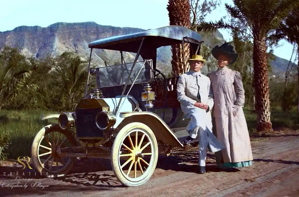 A colorized vintage photograph shows a man and woman posing beside a classic early 1900s Ford Model T car with spoked wheels…