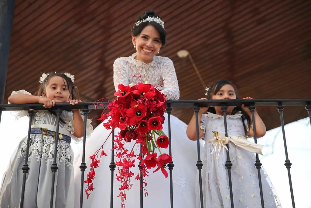 A smiling bride in a white lace dress and tiara stands on a balcony holding a large red bouquet, flanked by two young girls…
