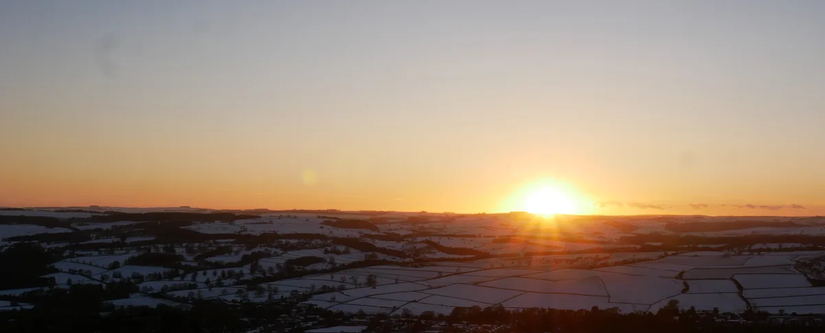 A panoramic view of a sunrise over a vast, snow-covered rural landscape with patchwork fields and distant hills, the sun…