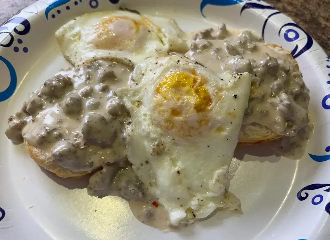 A close-up photo of a plate of breakfast food featuring two sunny-side-up eggs and three biscuits generously covered in…