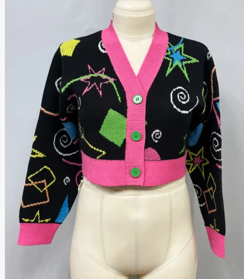 A black, cropped, v-neck cardigan with bright pink trim and green buttons is displayed on a mannequin. The sweater features…