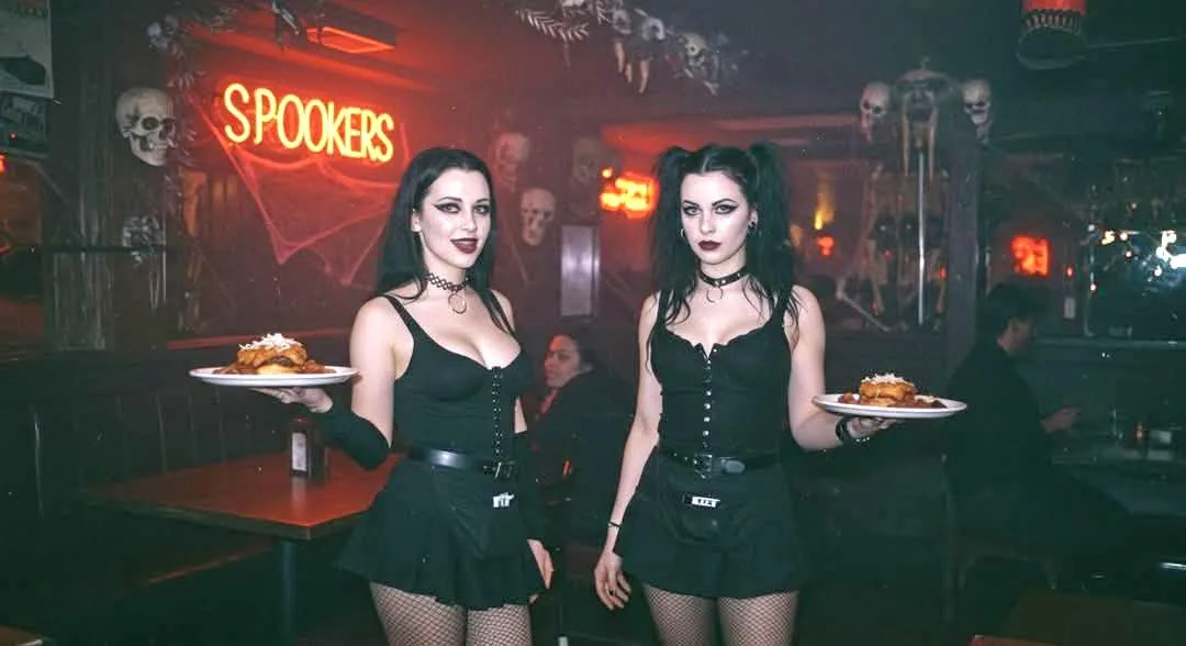 Two women in gothic attire, including black corsets, skirts, fishnet stockings, and chokers, stand in a dimly lit restaurant…