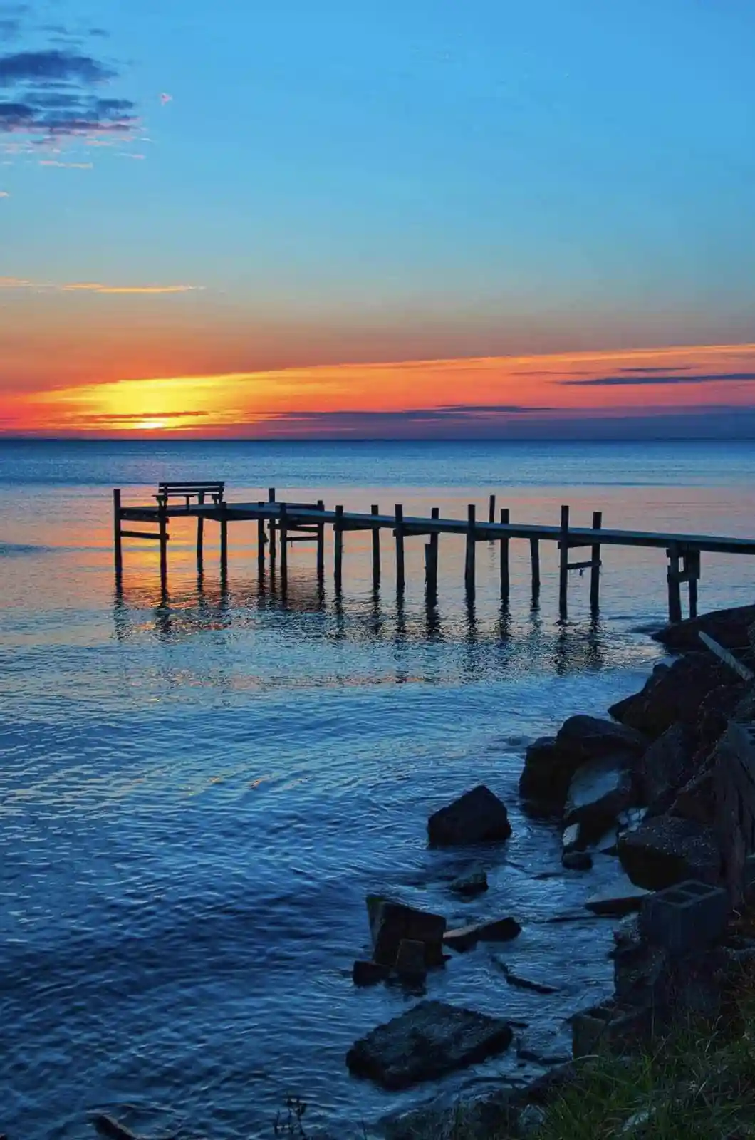 A serene sunset over a calm body of water with a wooden pier extending into the distance. The sky displays vibrant orange…