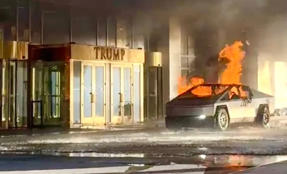 A Tesla Cybertruck is on fire next to a building with a prominent 'TRUMP' sign above its entrance. The vehicle is engulfed…