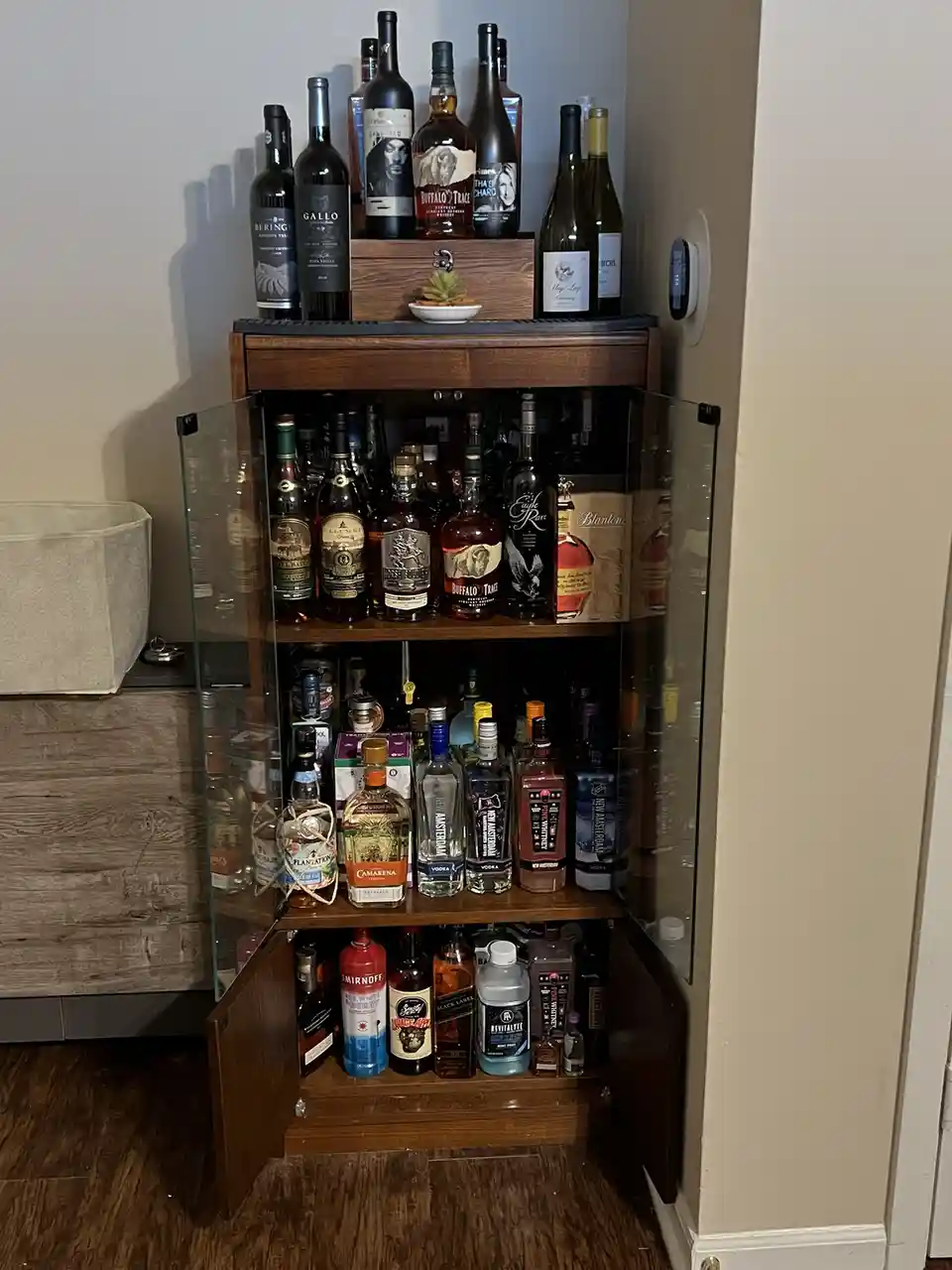 A wooden, glass-fronted mini-bar cabinet with its doors open, revealing multiple shelves packed with various liquor bottles…