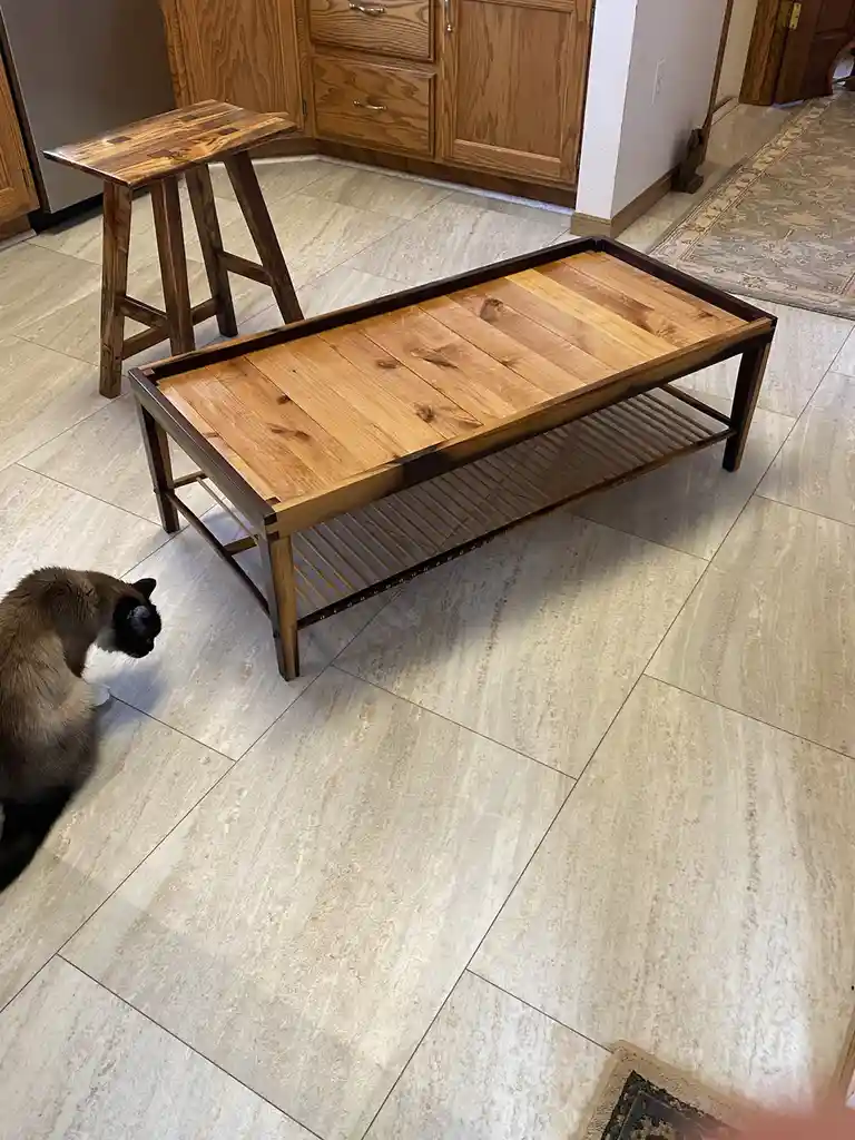 A siamese cat is seen from the side, looking down at the floor near a wooden coffee table with a lower shelf, in a room with…
