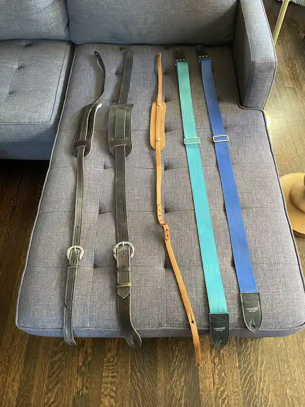 Five guitar straps of varying colors and materials — two dark brown leather, one tan leather, one teal fabric, and one blue…