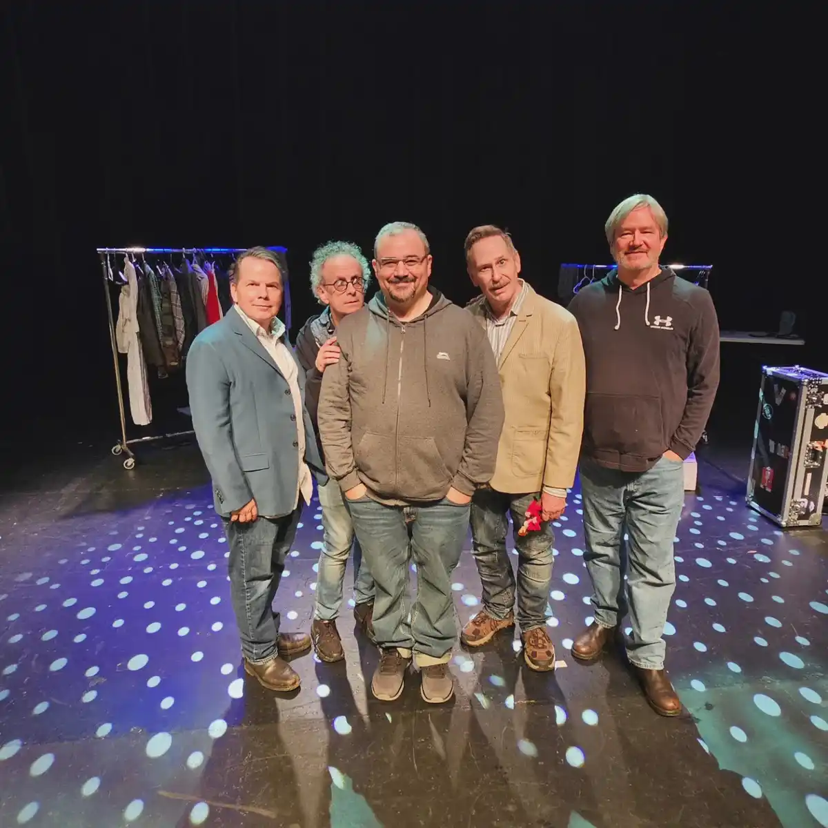 Five men stand together on a stage with a dark backdrop and a clothing rack visible behind them. The stage floor is…