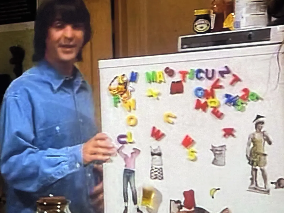 A man in a blue shirt stands beside a refrigerator covered in colorful magnetic letters and various fridge magnets…