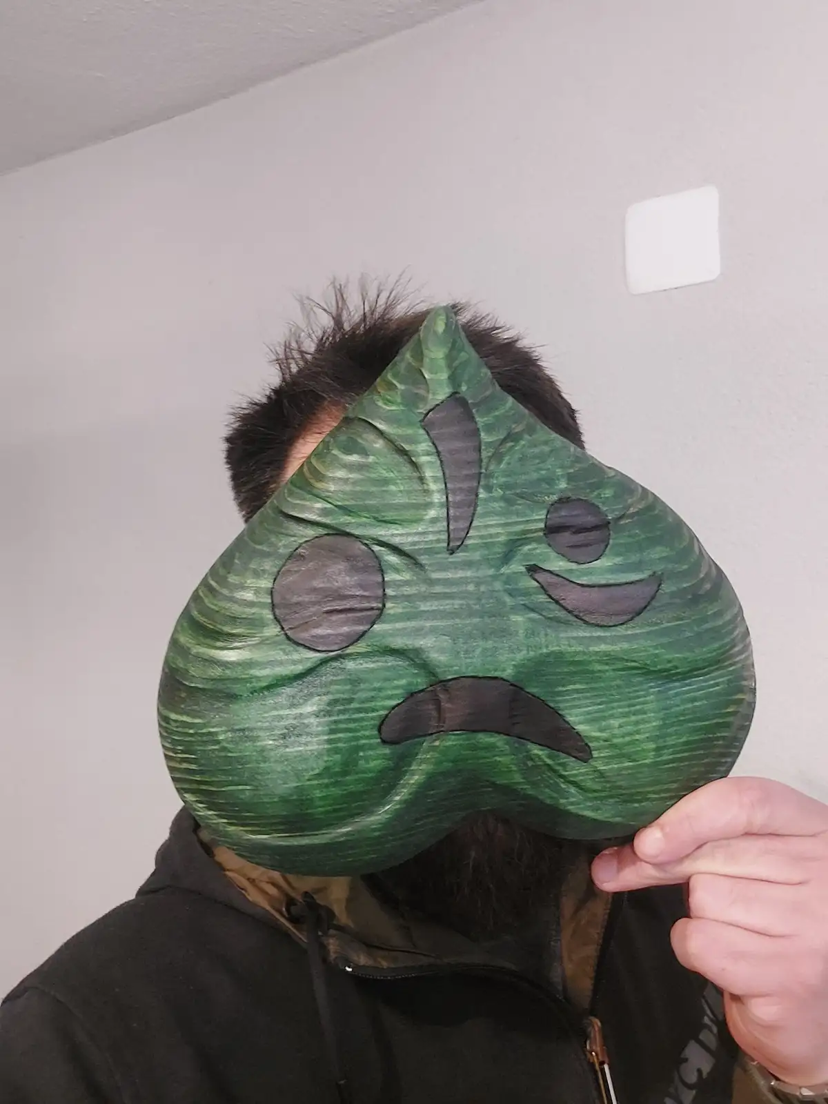 A person with dark hair and a beard wears a large, green, leaf-shaped mask with cartoonish black eyes, nose, and mouth. The…