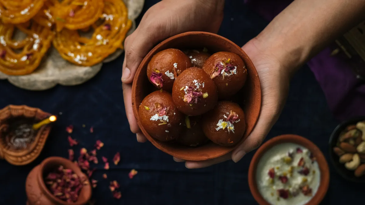 A pair of hands holds a small clay bowl filled with round, brown Indian sweets (laddus) topped with rose petals and silver…