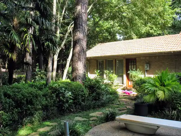 A tranquil suburban backyard with a brick house, lush greenery, and a stone path leading to the front door. In the…