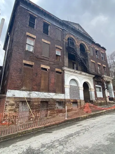 a dilapidated, multi-story brick building with boarded-up windows, broken archways, and visible decay under an overcast sky…