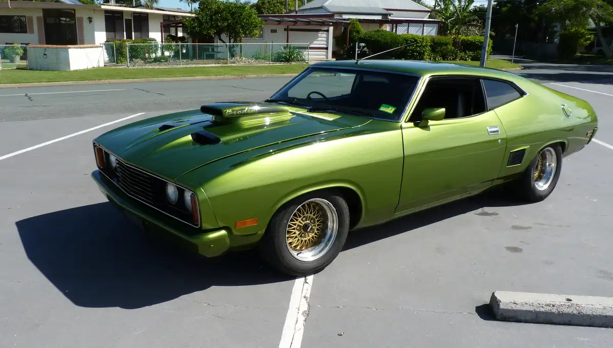 A shiny, lime-green classic muscle car with gold wheels and a hood scoop is parked in a sunny parking lot, with the name…