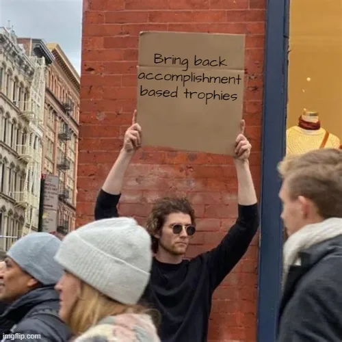 A man with curly hair and sunglasses holds up a cardboard sign that reads 'Bring back accomplishment based trophies' while…