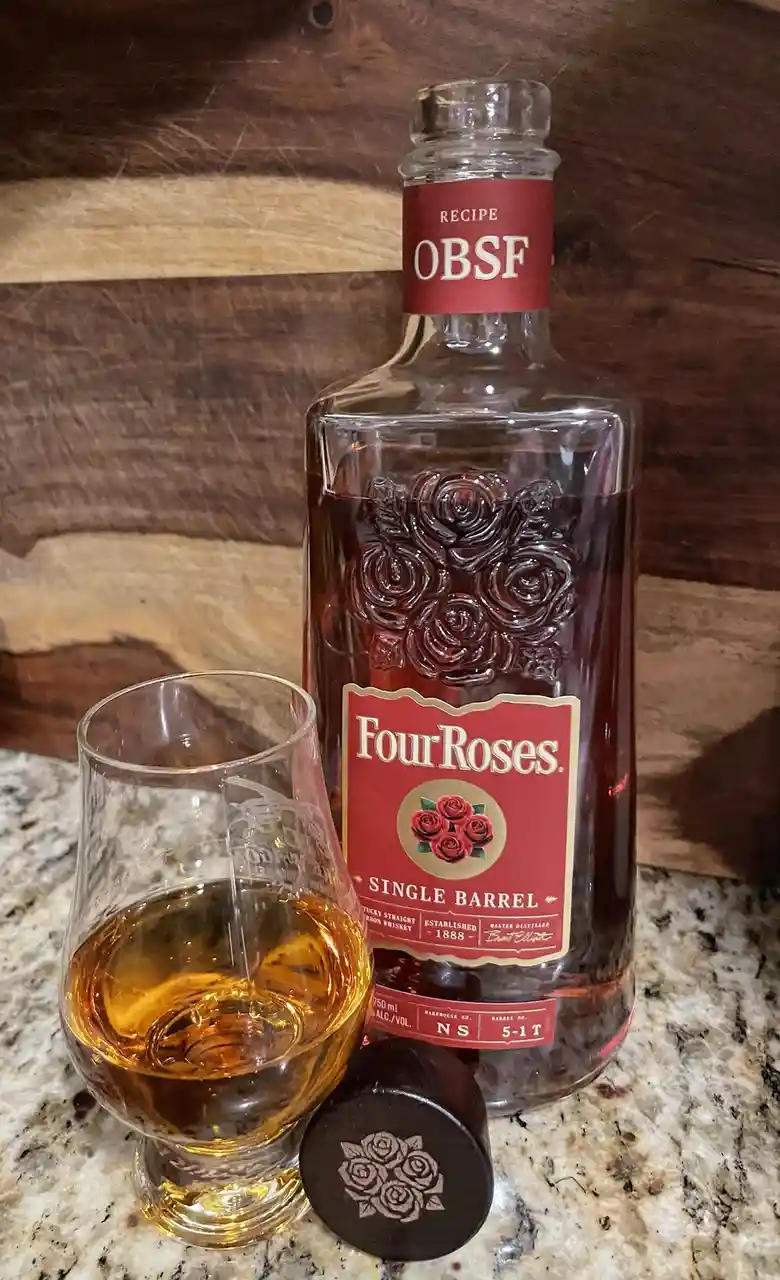 A bottle of Four Roses Single Barrel bourbon whiskey with its cap off sits next to a glass containing amber liquid, both…