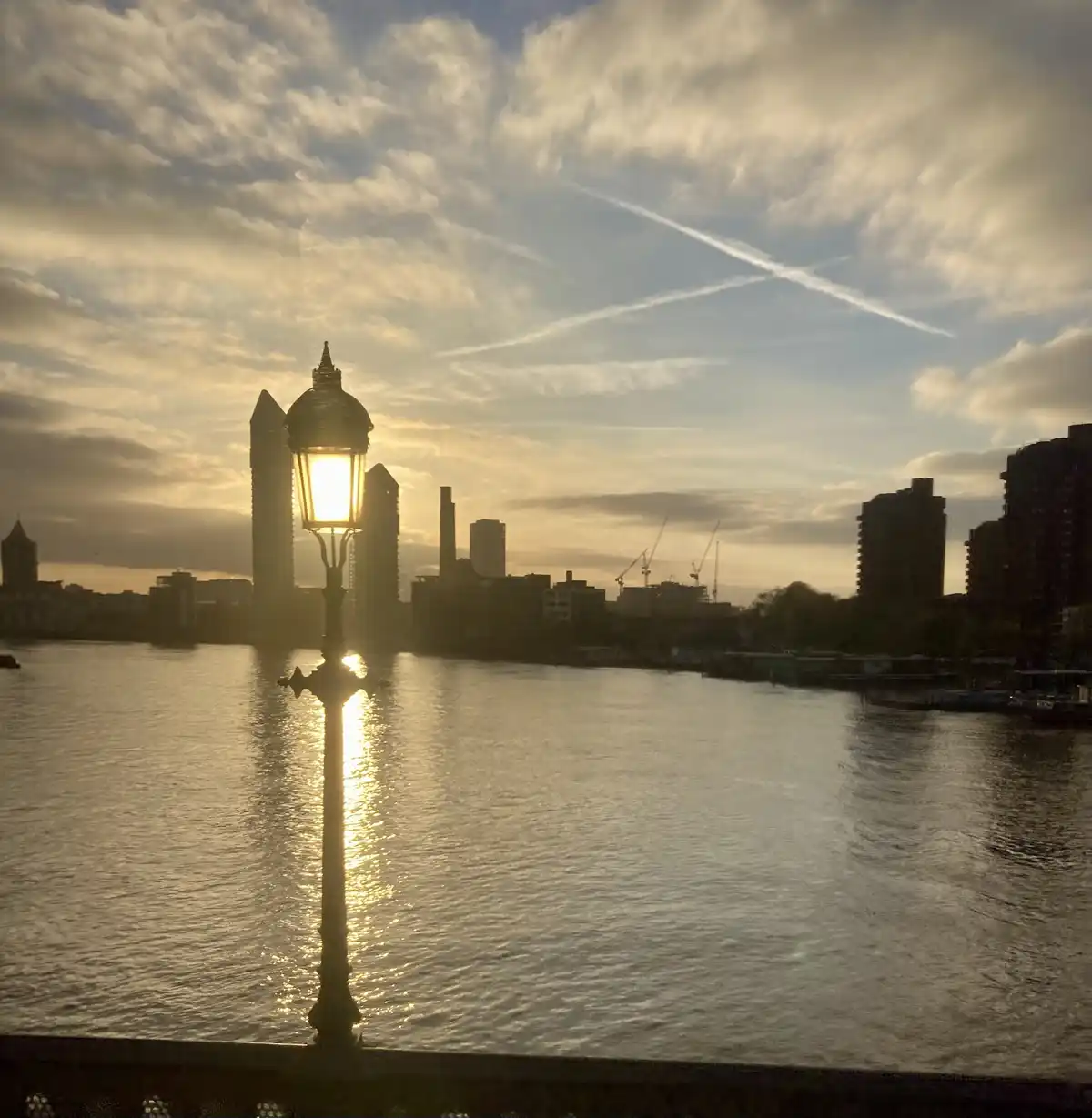 A serene sunset photo of a river with a lit ornate streetlamp in the foreground, its light reflecting on the water, with a…