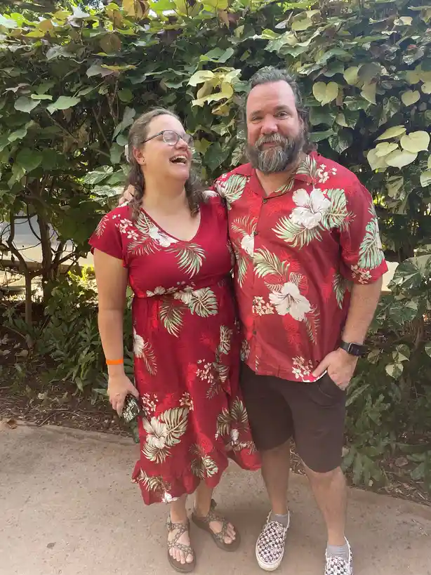 A man and woman stand together outdoors in front of lush green foliage, both wearing matching red shirts with white and…