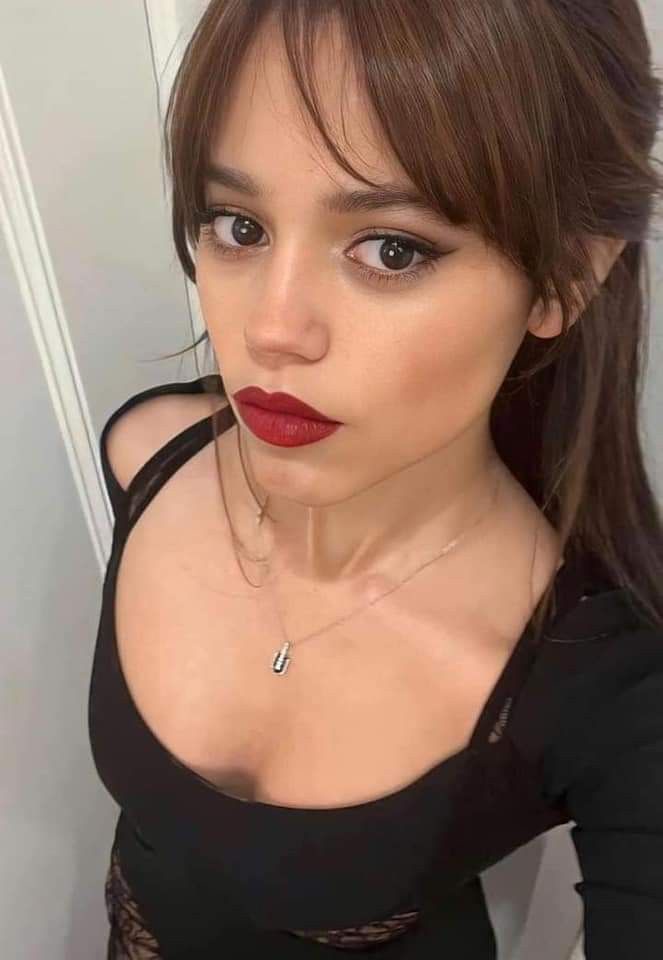 A woman with brown hair and red lipstick stares directly at the camera with a skeptical expression while wearing a black top…