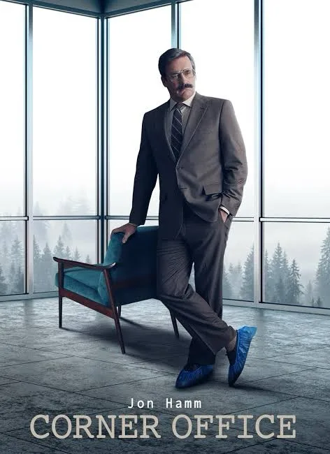 A man in a brown suit and blue shoes leans casually against a teal chair in a modern corner office with large windows…