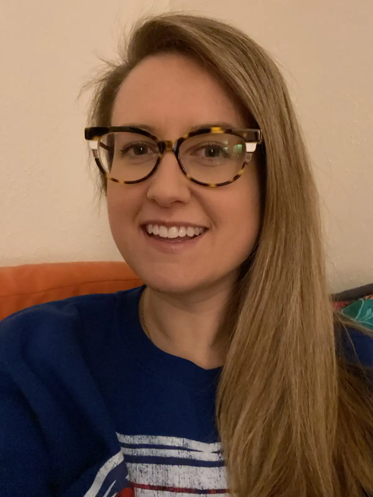 A woman with long blonde hair and tortoiseshell glasses smiles at the camera while wearing a blue shirt with white stripes…