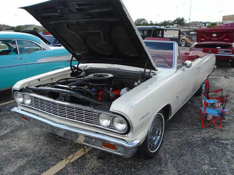 A white 1960s Chevrolet convertible with its hood open, revealing an engine bay that appears mostly empty or stripped down…