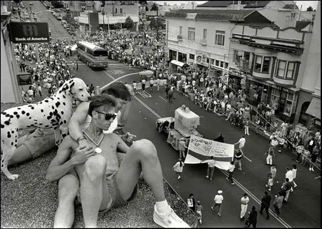 A black-and-white photo shows a man in sunglasses sitting on a curb with a large inflatable dalmatian beside him, while a…