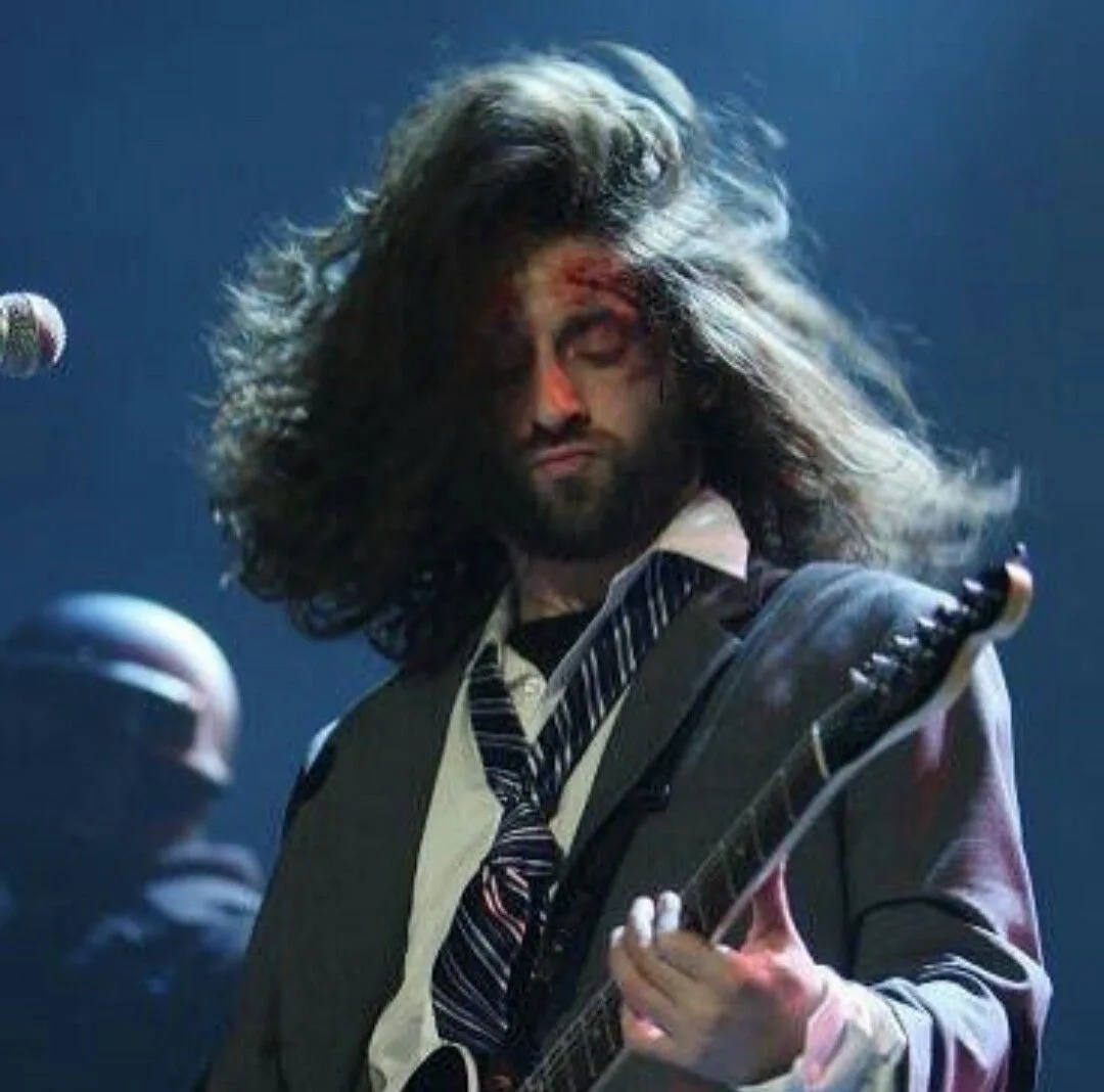 A man with long, wild hair and a beard, wearing a suit and striped tie, plays an electric guitar on stage with a bruised and…