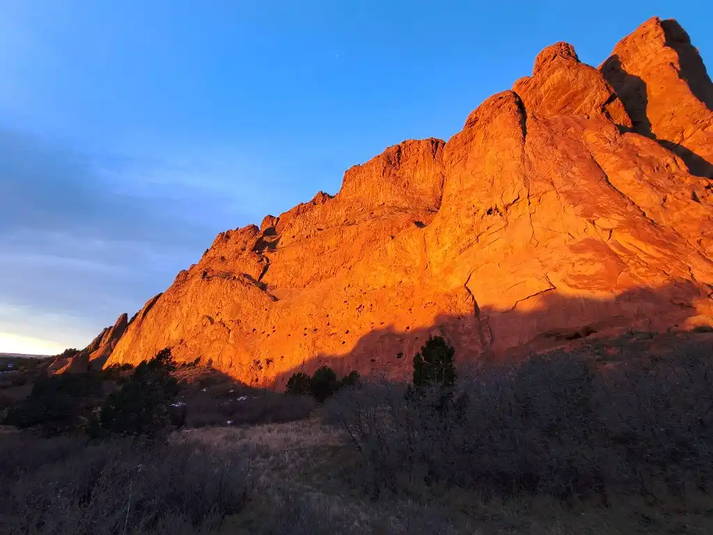 A scenic photo of red rock formations bathed in warm, golden sunlight against a clear blue sky, with dark silhouettes of…
