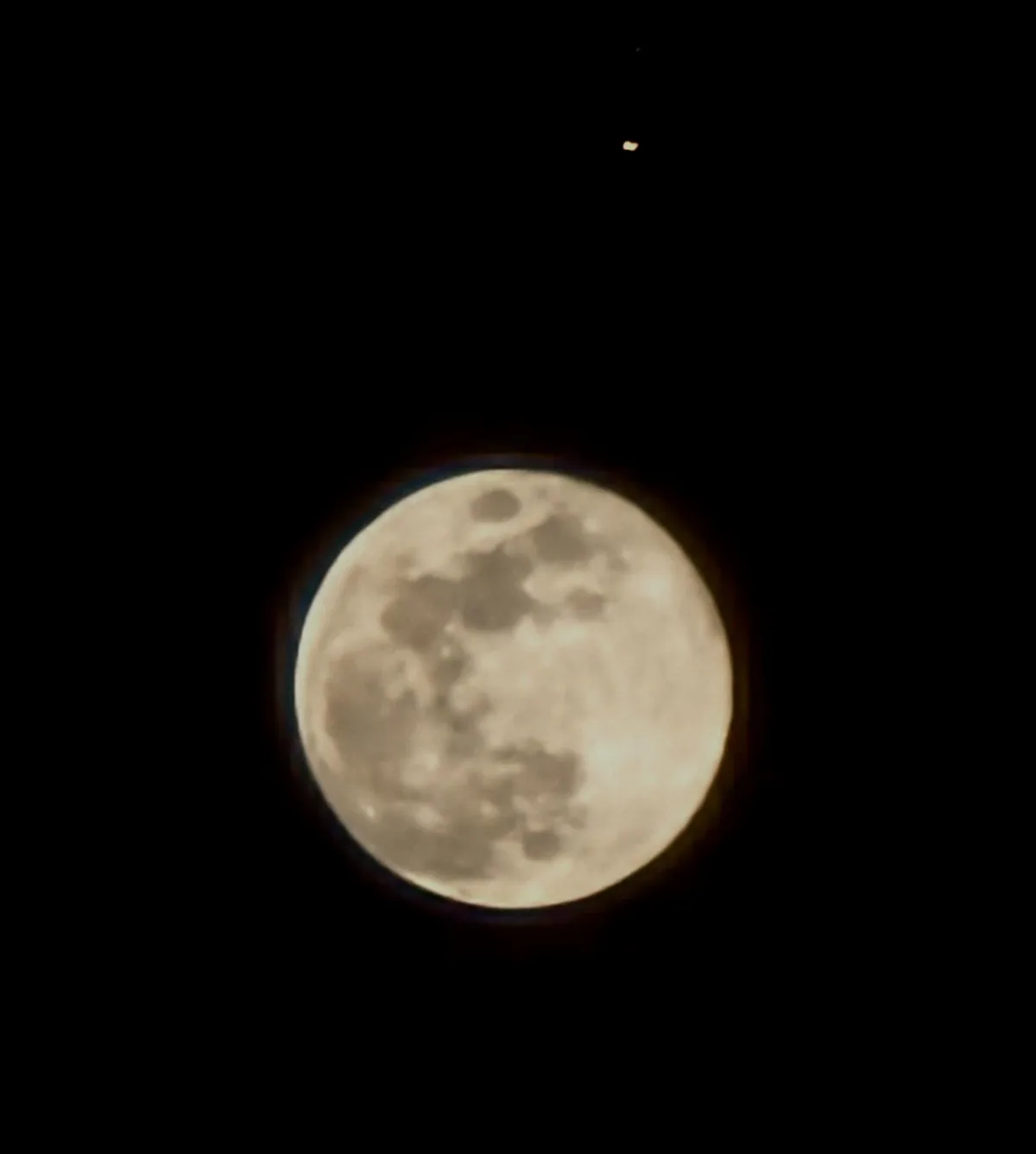 A close-up photo of a full moon against a black night sky, with a small distant light visible in the upper right corner. The…